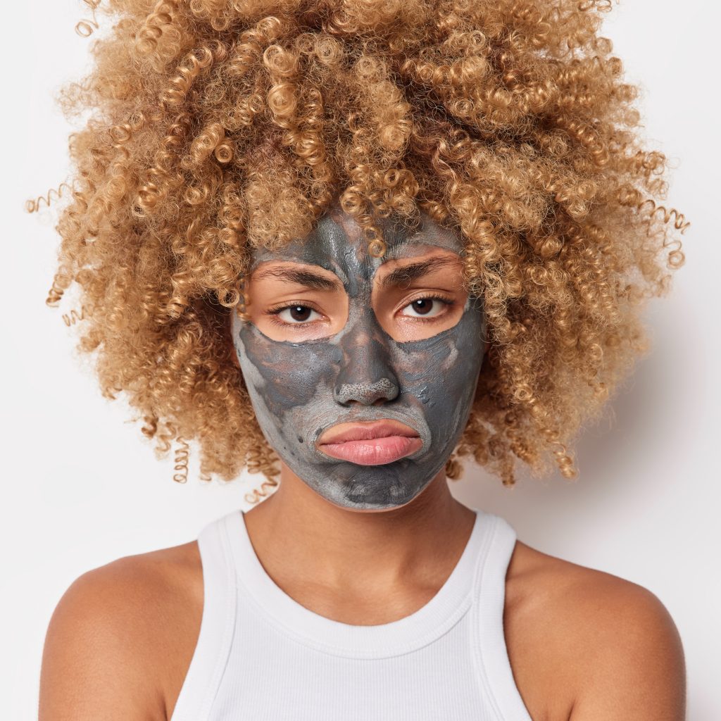 Portrait of upset curly haired young female model applies clay mask on face to remove pores undergoes daily beauty procedures wears t shirt isolated over white background. Facia care concept