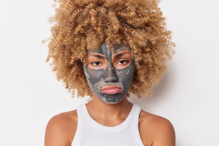 Portrait of upset curly haired young female model applies clay mask on face to remove pores undergoes daily beauty procedures wears t shirt isolated over white background. Facia care concept