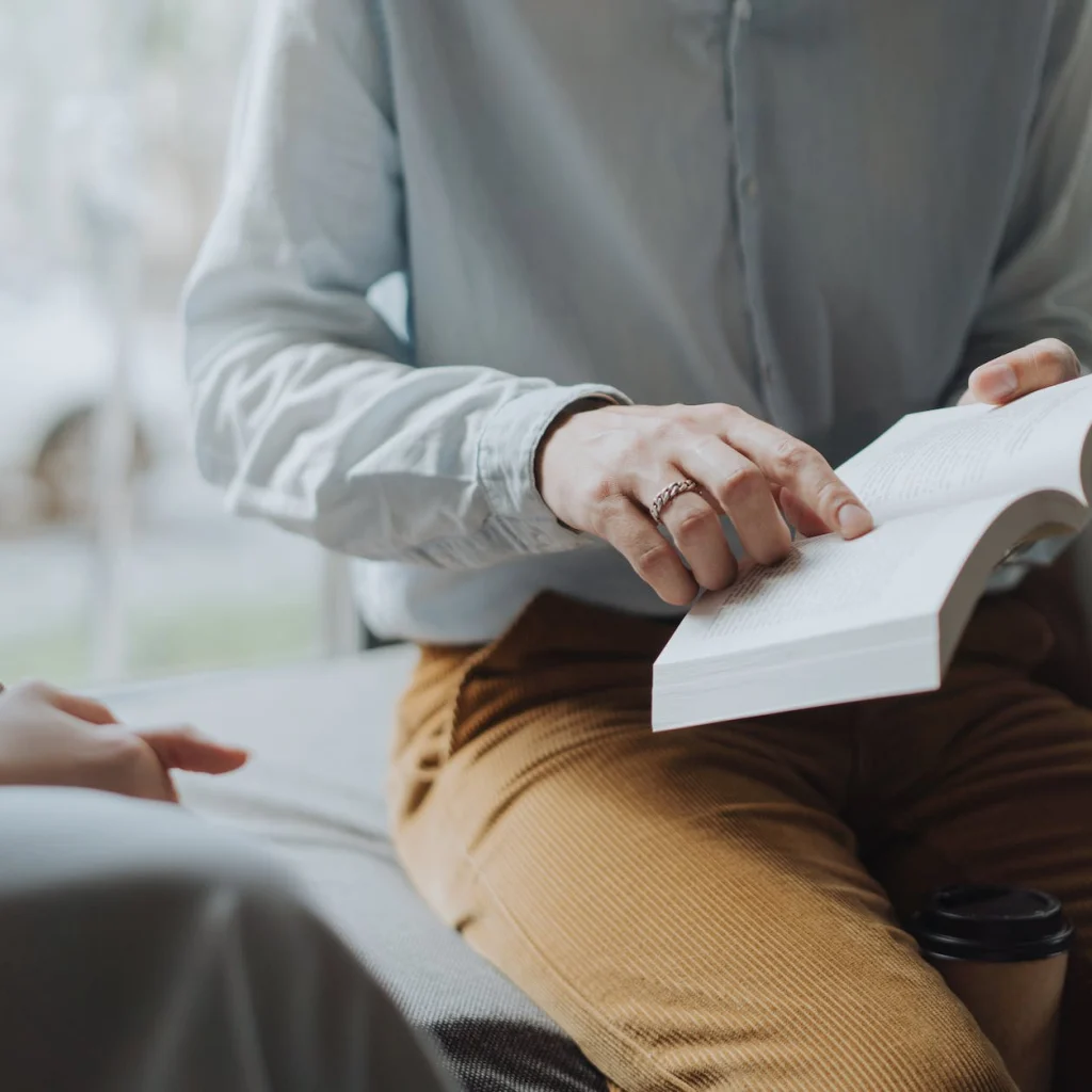 man in white dress shirt and brown pants sitting on white chair reading book
