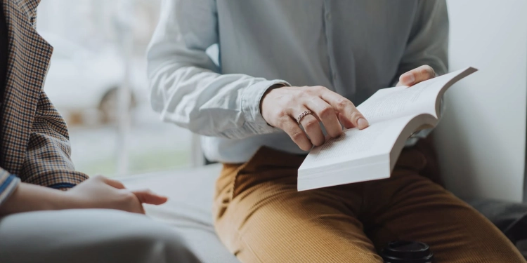 man in white dress shirt and brown pants sitting on white chair reading book