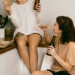 Happy female hipster enjoying slice of pizza and drinking beverage while talking to happy friend sitting with crossed legs on chair in apartment