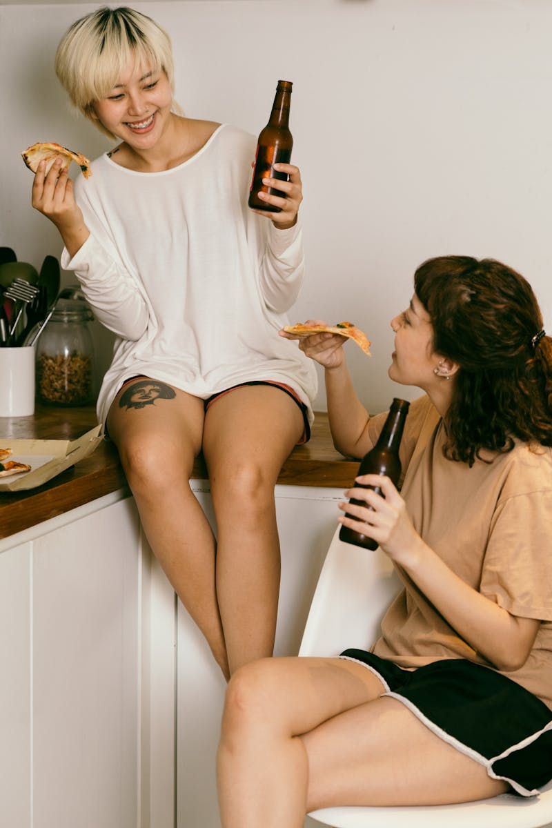 Happy female hipster enjoying slice of pizza and drinking beverage while talking to happy friend sitting with crossed legs on chair in apartment