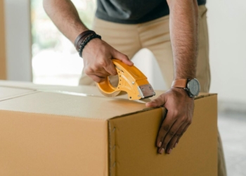 Crop faceless young male with wristwatch using adhesive tape while preparing cardboard box for transportation