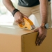 Crop faceless young male with wristwatch using adhesive tape while preparing cardboard box for transportation