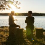 man in black jacket standing beside body of water during sunset