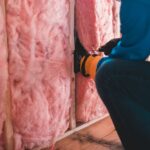 Crop unrecognizable worker in gloves sitting on haunches and insulating with pink stone wool