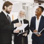 Three businessmen in suits discussing documents in a modern office setting, engaging in teamwork.