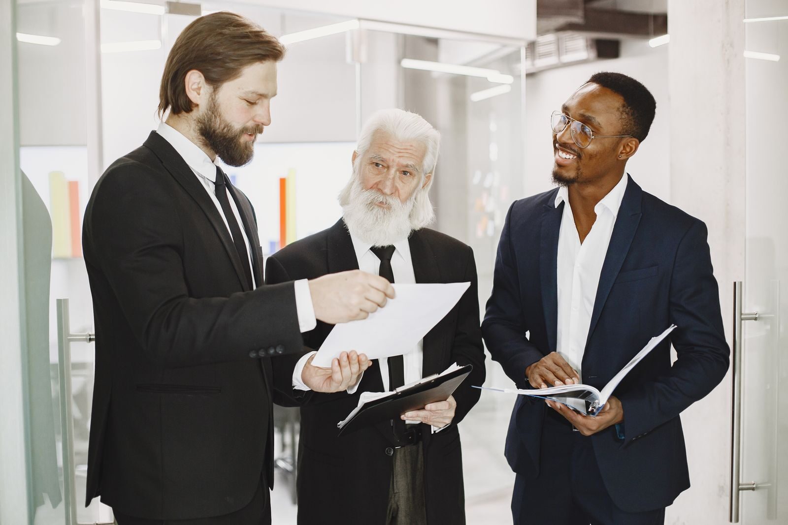 Three businessmen in suits discussing documents in a modern office setting, engaging in teamwork.