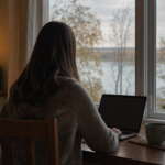 A woman sits at a kitchen table facing a window with a laptop open, a river visible through bare trees outside.